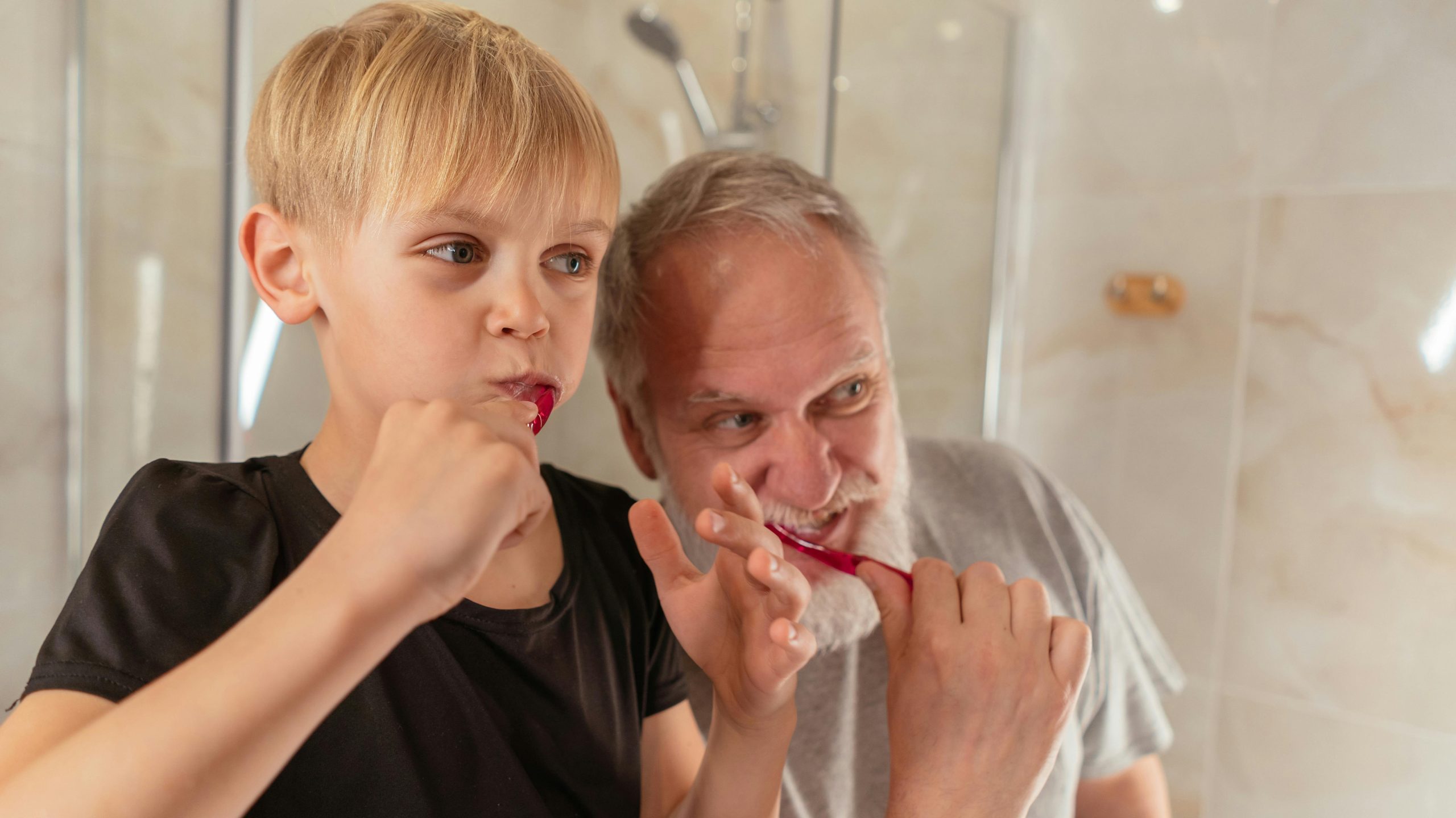 An elderly man and a young boy brushing their teeth together, promoting oral care for seniors. NYC Dentist post.